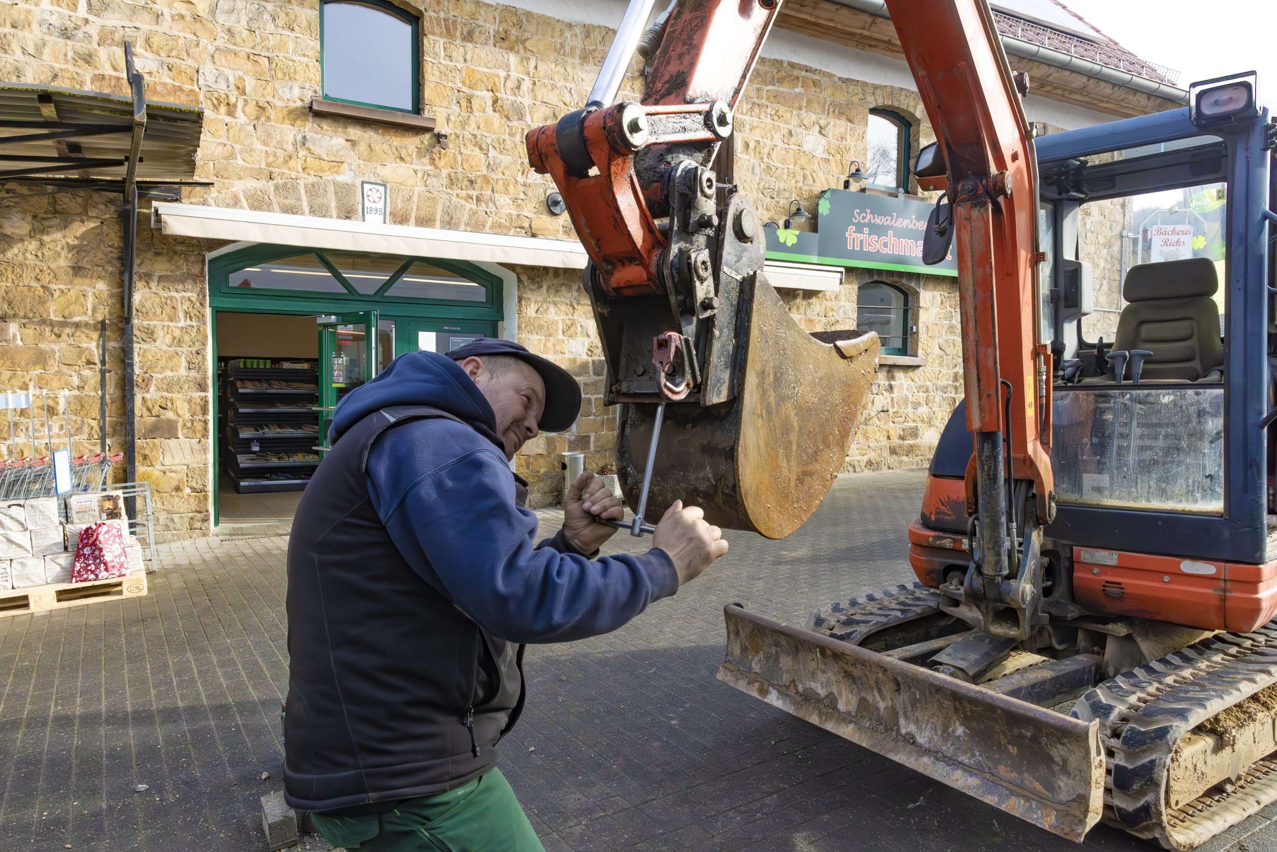 Ein Mann repariert mit einem Werkzeug den Löffel eines kleinen orangen Baggers vor einem Gebäude aus Sandstein mit grüner Tür und Schaufenster.