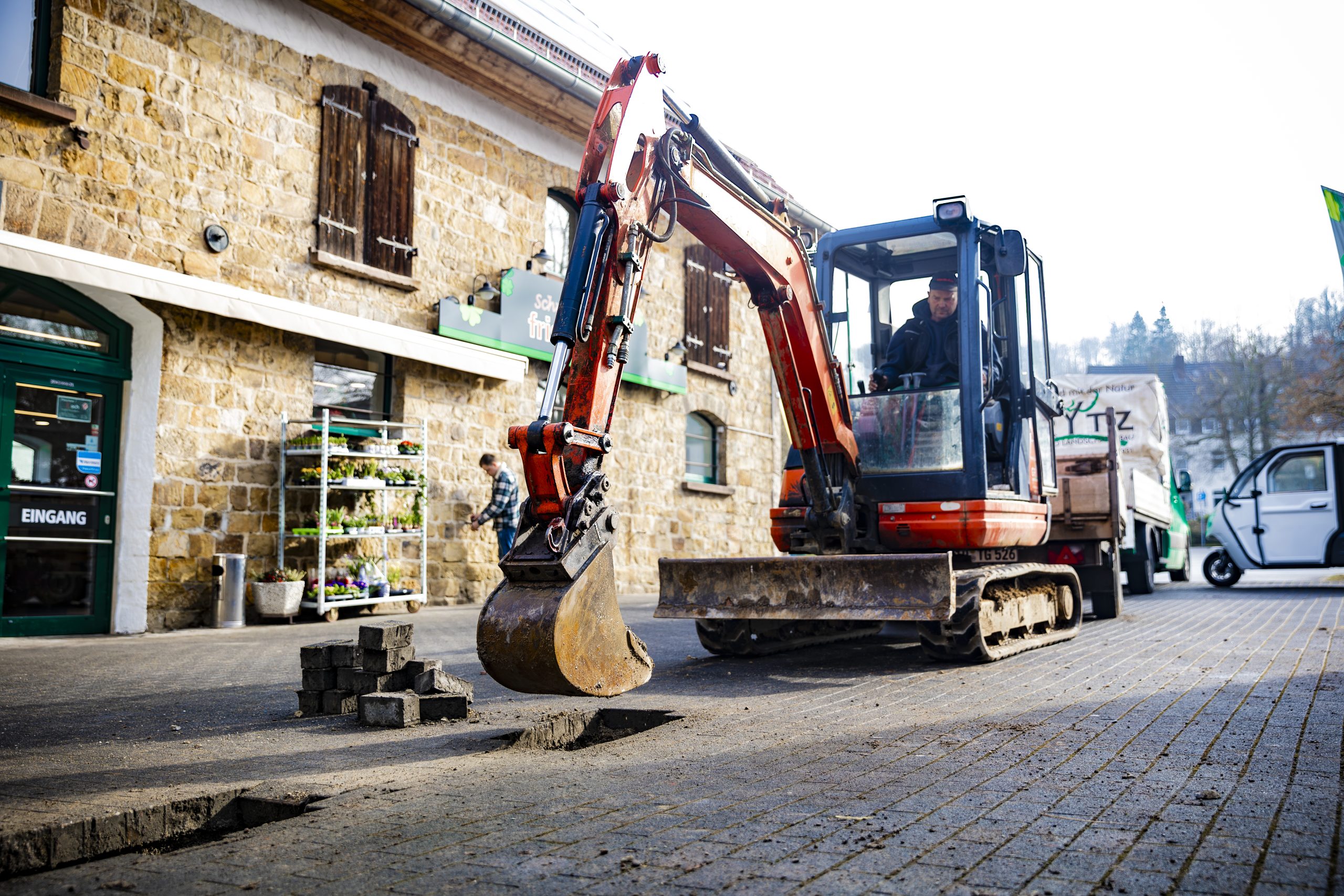 Kleiner Bagger mit rotem Arm, der auf einem gepflasterten Gehweg arbeitet, neben einem Steinmauergebäude.
