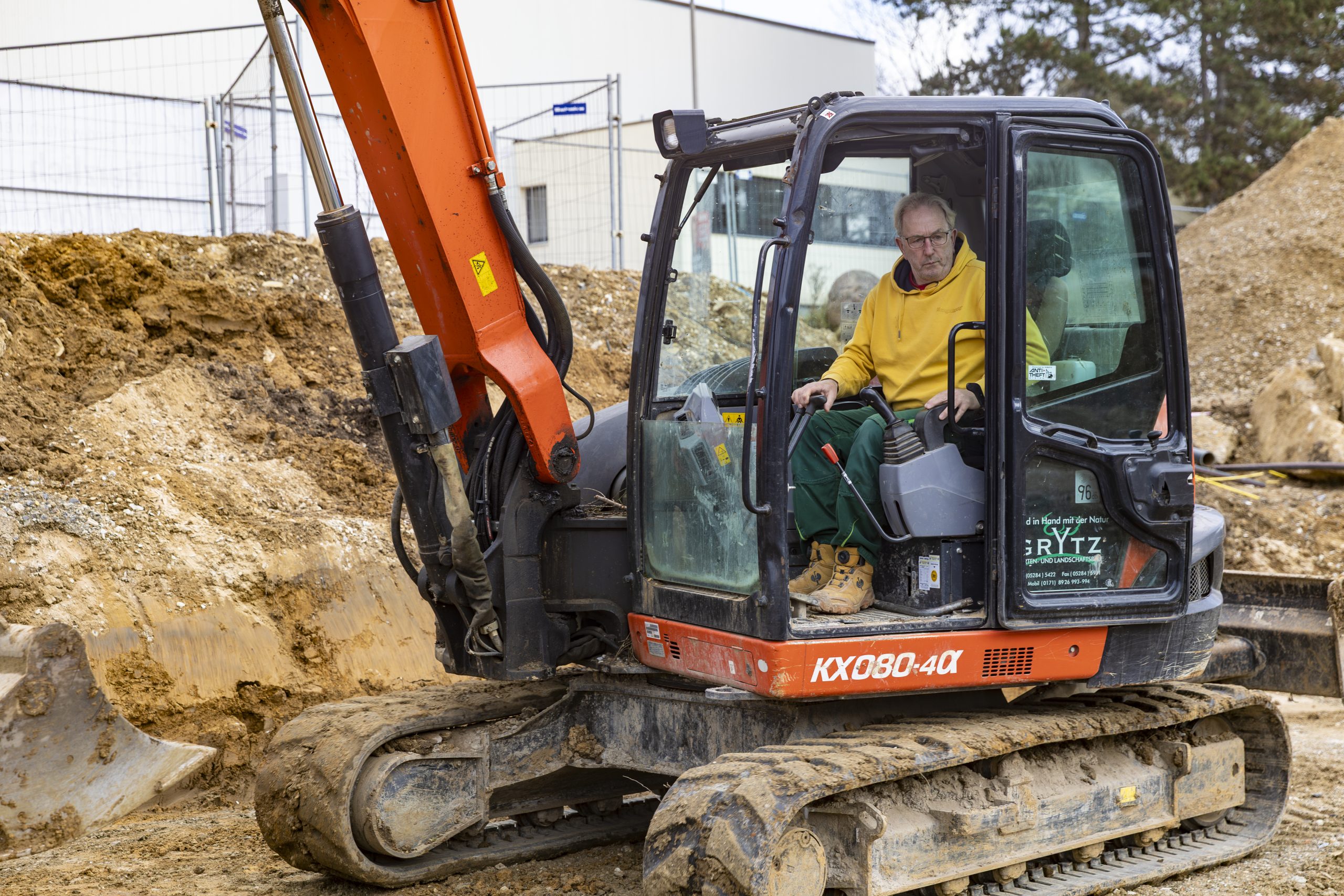 Mann in gelber Kapuzenjacke und grüner Arbeitskleidung sitzt im Führerhaus eines orangen Kettenbaggers auf einer Baustelle mit Erdhaufen im Hintergrund.