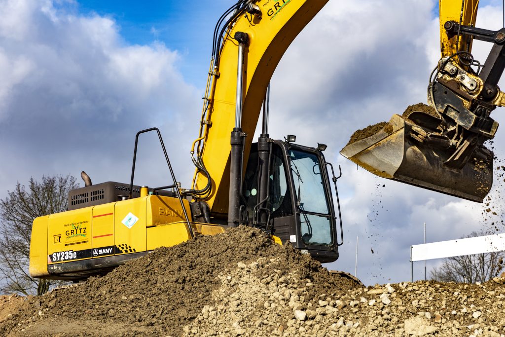 Gelber Bagger der Marke SANY beim Ausheben von Erde auf einer Baustelle unter bewölktem Himmel.