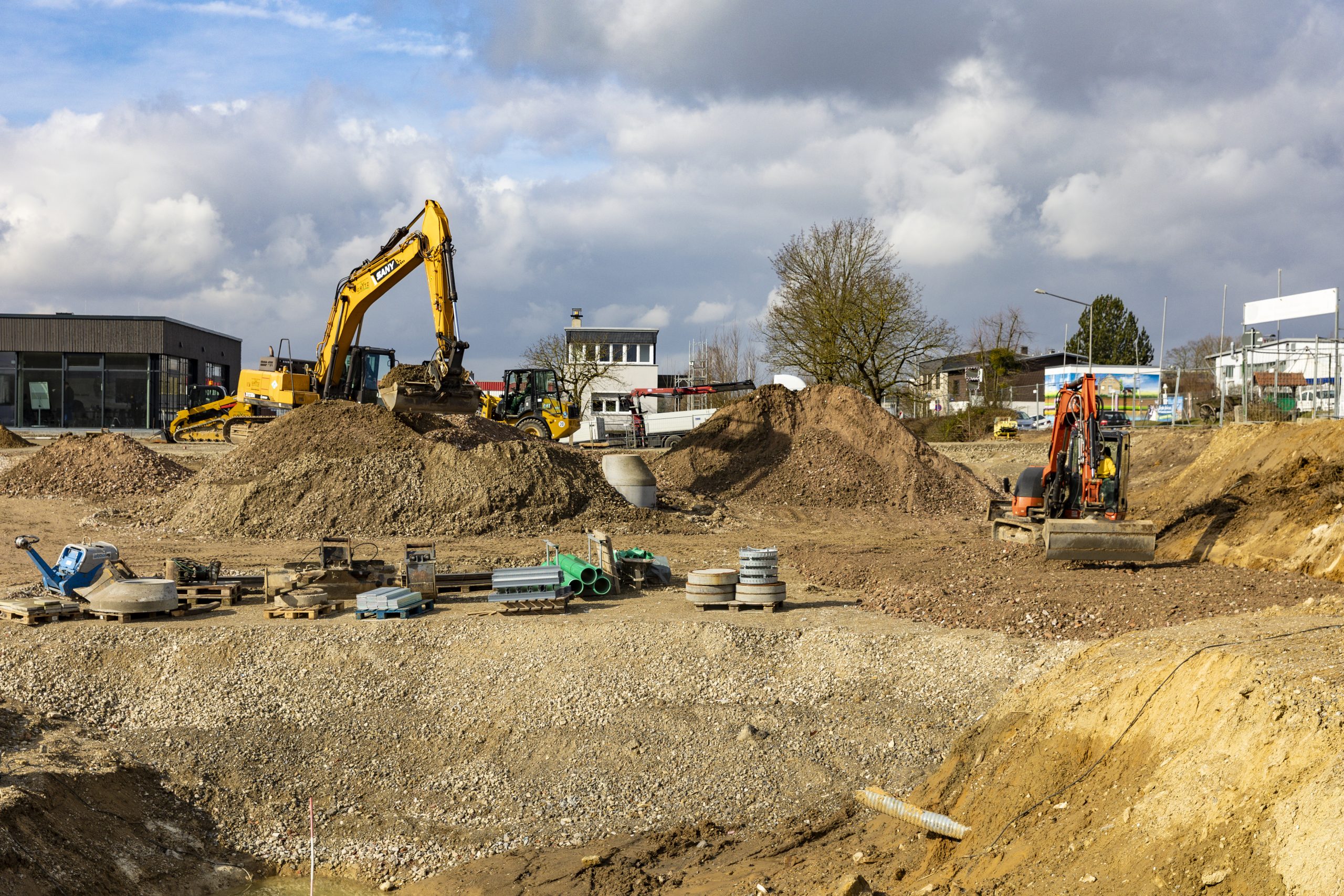 Baustelle mit großen Erdbergen, mehreren Baggern und Baumaschinen, sowie Baumaterialien auf Paletten unter bewölktem Himmel.