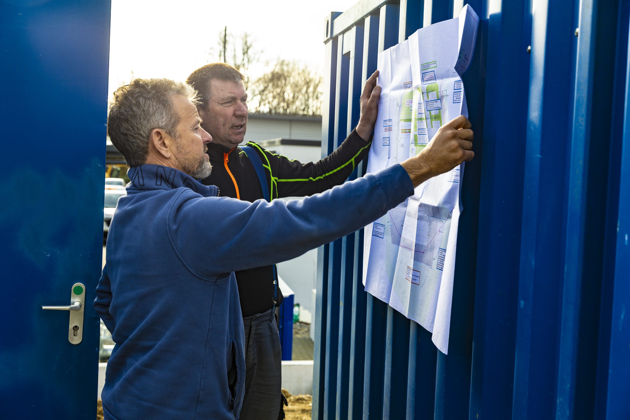 Thomas Grytz und sein Vorarbeiter betrachten gemeinsam eine Bauzeichnung, der an einer blauen Containerwand befestigt ist, im Freien bei Tageslicht.