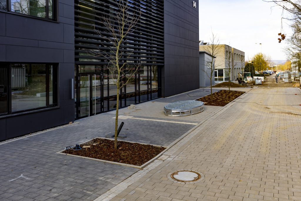 Moderne Gebäude mit schwarzer Fassade und großflächigen Fenstern, davor gepflasterter Gehweg mit jungen, kahlen Bäumen in quadratischen Erdbeeten, weiter hinten Baustelle mit Baufahrzeugen und Paletten auf breiter Straße