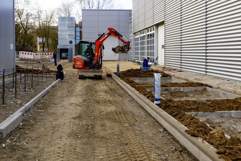 Baustelle zwischen zwei modernen Industriegebäuden mit Bagger und Bauarbeitern, die Erdarbeiten und Vorbereitungen für Pflasterungen durchführen.