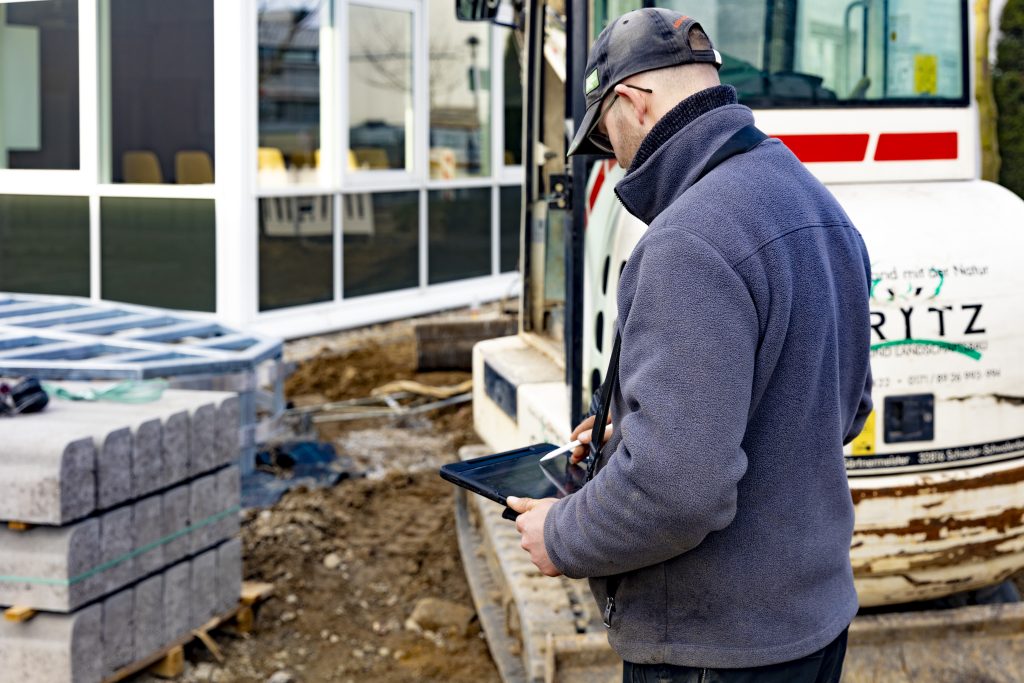 Der Vorarbeiter bedient mit einem Stift ein Tablet auf einer Baustelle, im Hintergrund Baumaschine und Baumaterialien.
